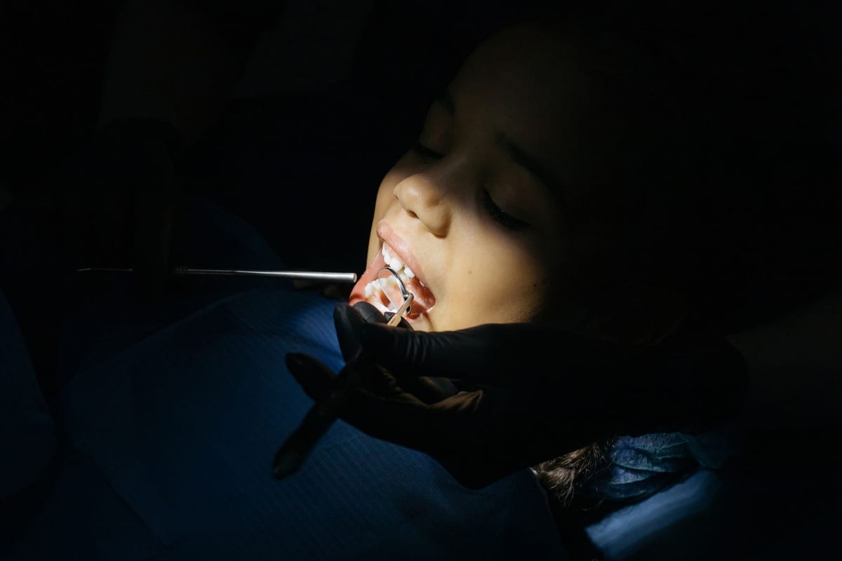 A young girl reclined in the dental chair while a gloved hand holds a mirror at her lips